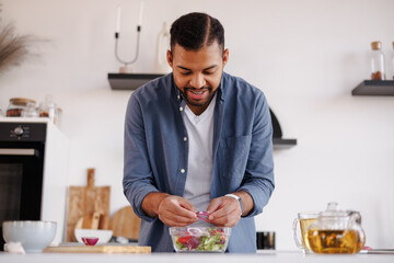 Smiling african american man adding onion in fresh salad near tea in kitchen at home