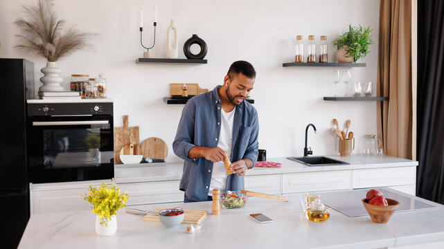 African american man in earphones seasoning fresh salad near tea and smartphone in kitchen