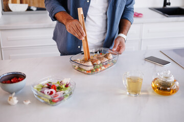 African american man holding wooden tongs near food in baking dish, fresh salad and herbal tea in kitchen
