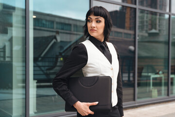 Young businesswoman in a vest holds a briefcase while standing confidently outside an office building on a sunny day