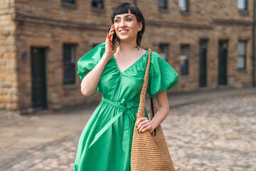 Fototapeta premium Smiling woman in a vibrant green dress walking and talking on her phone on a cobblestone street in a quaint urban setting
