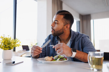 African american man eating fresh salad and chicken meat near tea and smartphone at home