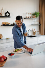 Positive african american man holding herbal tea and using laptop near dinner in kitchen