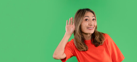 A cheerful woman in an orange shirt playfully listening with one hand cupped to her ear against a...