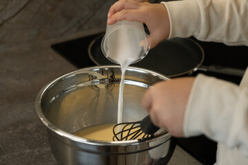 A small hands holds a whisk and a measuring cup and pours milk into a bowl