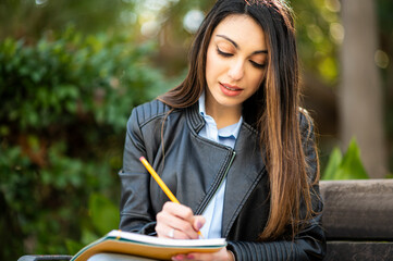 Beautiful female college student reading a book on a bench in a park and taking notes