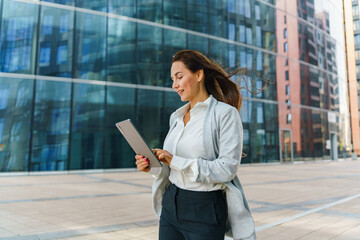 Young Professional Woman Walks Outside a Modern Urban Building While Using a Tablet on a Sunny Day