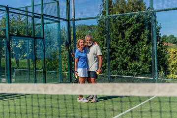 Senior elderly padel player couple embracing on court with paddles