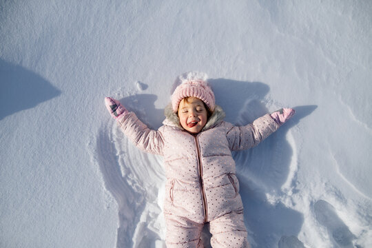 Portrait of small girl making snow angel, enjoying winter holiday in the mountains with family. Girl lying and playing in snow.