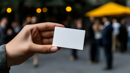A person holds a blank card while attending a networking event outdoors, surrounded by groups of professionals engaging in conversation during an evening gathering