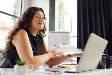 In a bright office, a plus-size woman with long curly hair smiles contentedly while working on her laptop.