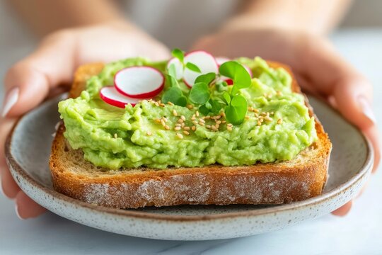  Tostada de pan integral con pur&eacute; de aguacate, rodajas de r&aacute;bano y microverdes, sostenida por unas manos.


