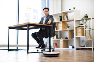Asian male adult working in modern office while multitasking with robot vacuum cleaner....