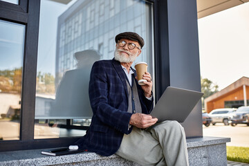A fashionable senior man sips coffee, working on his laptop, dressed in a checkered blazer and flat...