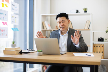 Asian businessman in formal attire engaging in online video call from contemporary office. Mid-adult male raising hands, possibly for emphasis or clarification, while working on laptop.