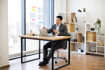Asian male business professional in modern office setting working on laptop during video conference. Bright, organized workspace with shelves, plants, and city view through large window.