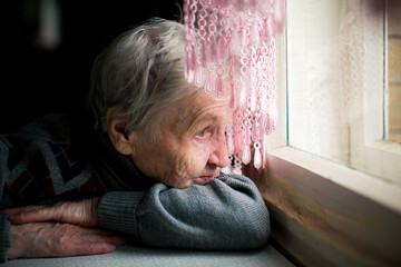 An elderly woman looks sadly out the window of her home, her eyes reflecting a deep sense of...