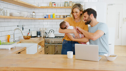 Busy Family In Kitchen At Breakfast With Parents Working On Laptop And Caring For Baby Son