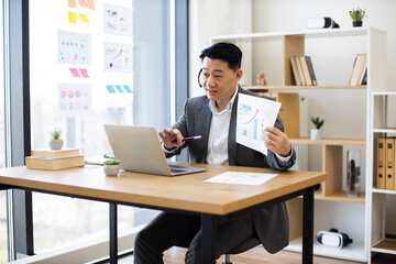 Asian male businessman in modern office using laptop and wearing headset during an online video call. He holds documents with graphs and charts, presenting financial report or analysis.