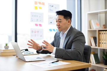 Asian businessman in professional setting engaged in online video call explaining business concepts. Confident, middle-aged male in formal attire speaking with expressive hand gestures.