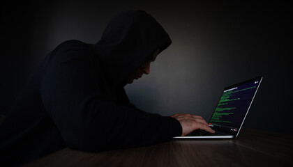 A hooded individual is typing on a laptop, with code displayed on the screen, set in a dimly lit...