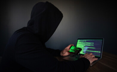 A hooded person operates a laptop and smartphone displaying green code, symbolizing hacking,...