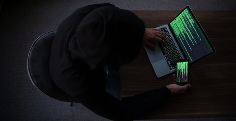 Top view on hooded figure sits at desk using laptop and smartphone, both displaying green code. The...