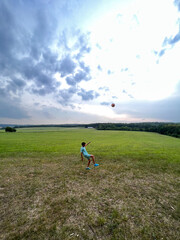 Child playing soccer in a vast green field under cloudy sky