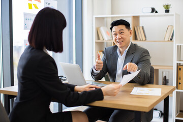 Asian businessman discusses report with Caucasian businesswoman in modern office. Professional setting includes laptops, charts, and documents.