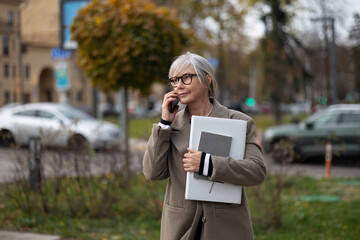 Senior businesswoman walking outside while talking on her phone, holding a laptop and documents on a city street