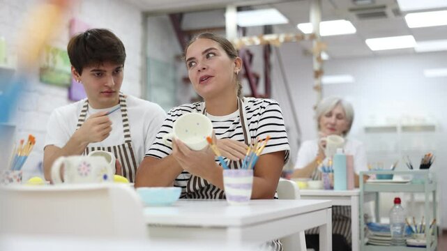 Cheerful young couple, guy and girl, actively engaged in creative hobby, decorating ceramic pottery together at table in cozy workshop setting