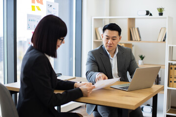 Asian businessman in grey suit reviews report with Caucasian businesswoman in a modern office. They are engaged in serious discussion, emphasizing teamwork and corporate environment.