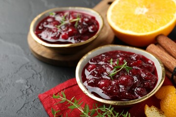 Tasty cranberry sauce in bowls, orange, thyme and cinnamon on black table, closeup