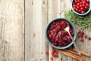 Tasty cranberry sauce in bowl, berries, rosemary and cinnamon sticks on wooden table, flat lay. Space for text