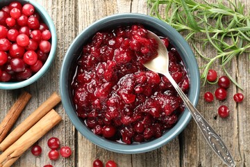 Tasty cranberry sauce in bowl, berries, rosemary and cinnamon sticks on wooden table, flat lay