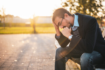 Depressed businessman in coat with eyeglasses sitting on the bench in park on a sunny autumn afternoon.