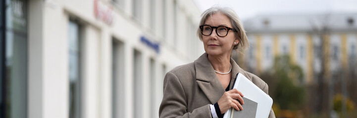 Elderly businesswoman walking confidently outside a modern office building on a crisp autumn day while holding a laptop