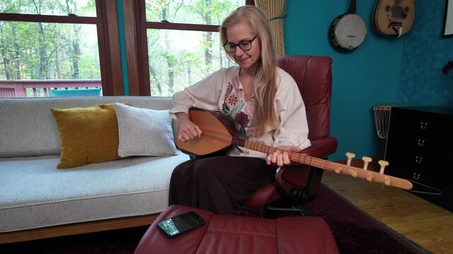 Mature woman with long blonde hair, wearing glasses and a white blouse with floral embroidery, sits in a red leather recliner playing a saz. The room has a teal wall and couch