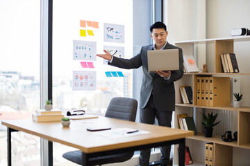 Naklejka premium Asian businessman presents data charts on laptop during online video call. He stands in office near panoramic window, analyzing business graphs.