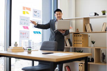 Naklejka premium Asian male businessman in modern office engaged in video call on laptop. Presenting data analysis with charts on panoramic window, creating professional engaging atmosphere.