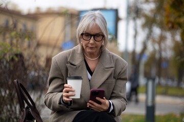 Older woman sitting on a bench outdoors, sipping coffee and browsing on her smartphone during a cool afternoon