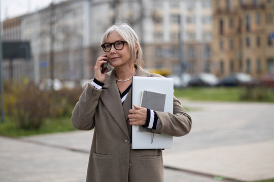 Senior businesswoman speaking on the phone while holding a notebook and documents on a city street in daylight