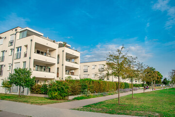 Modern apartment building surrounded by greenery under a clear blue sky. The residential complex features contemporary architecture with wooden accents, large windows, and balconies adorned with plant