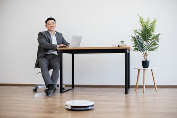 Asian businessman in suit works on laptop at modern office desk. Robot vacuum cleaner operates on wooden floor providing efficient cleaning. Bright workspace includes potted plant minimalist furniture