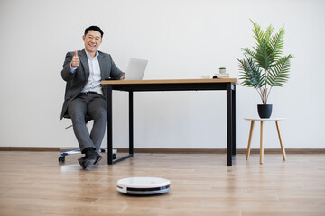 Asian male businessman gives thumbs up while working on laptop at desk against white wall. Robot vacuum cleaner operates on floor, highlighting modern technology integration in workspace.