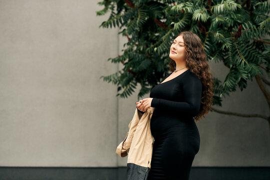 A young, beautiful plus-size woman walks outdoors in a black dress and beige trench coat amid autumn leaves.
