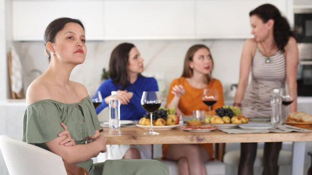 Upset young Asian woman sitting with crossed arms at table during home gathering while female friends expressing discontent and criticizing in background