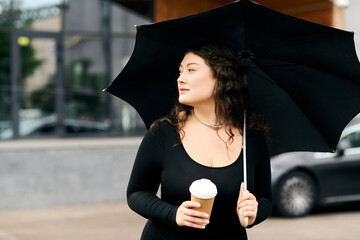 A young woman in a stylish black dress enjoys an autumn day, sipping coffee beneath her elegant...