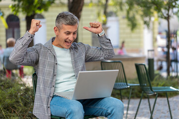 Happy Caucasian man freelancer working on laptop screams in triumph win celebrating good message news success game casino lottery money jackpot victory. Excited middle-aged guy on urban city street.