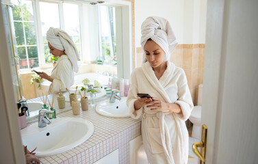 Attractive woman in a white bathrobe, towel wrapped around her head, stands in a bright bathroom while using her smartphone. The bathroom features a clean, modern design with natural light and decor
